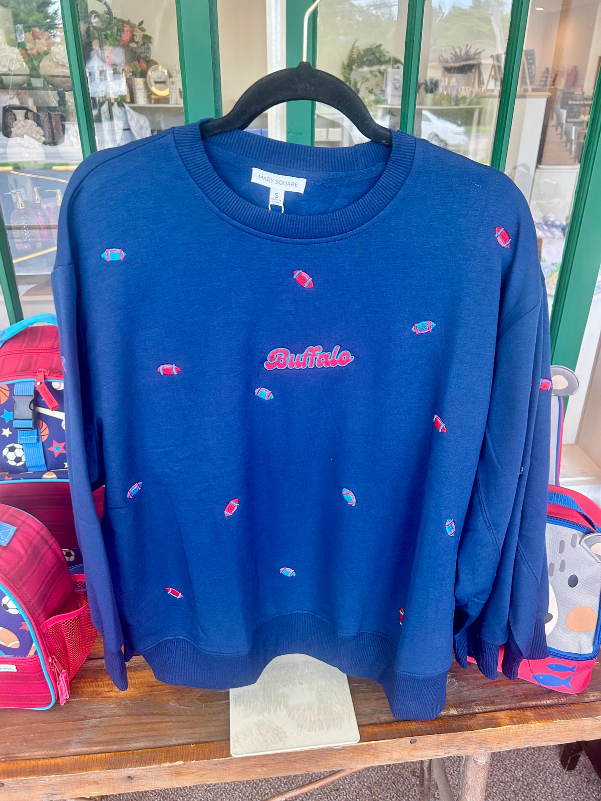 A navy blue crewneck sweatshirt with small embroidered fish motifs and 'Buffalo' text on the chest, hanging on a black hanger on a wooden table.
