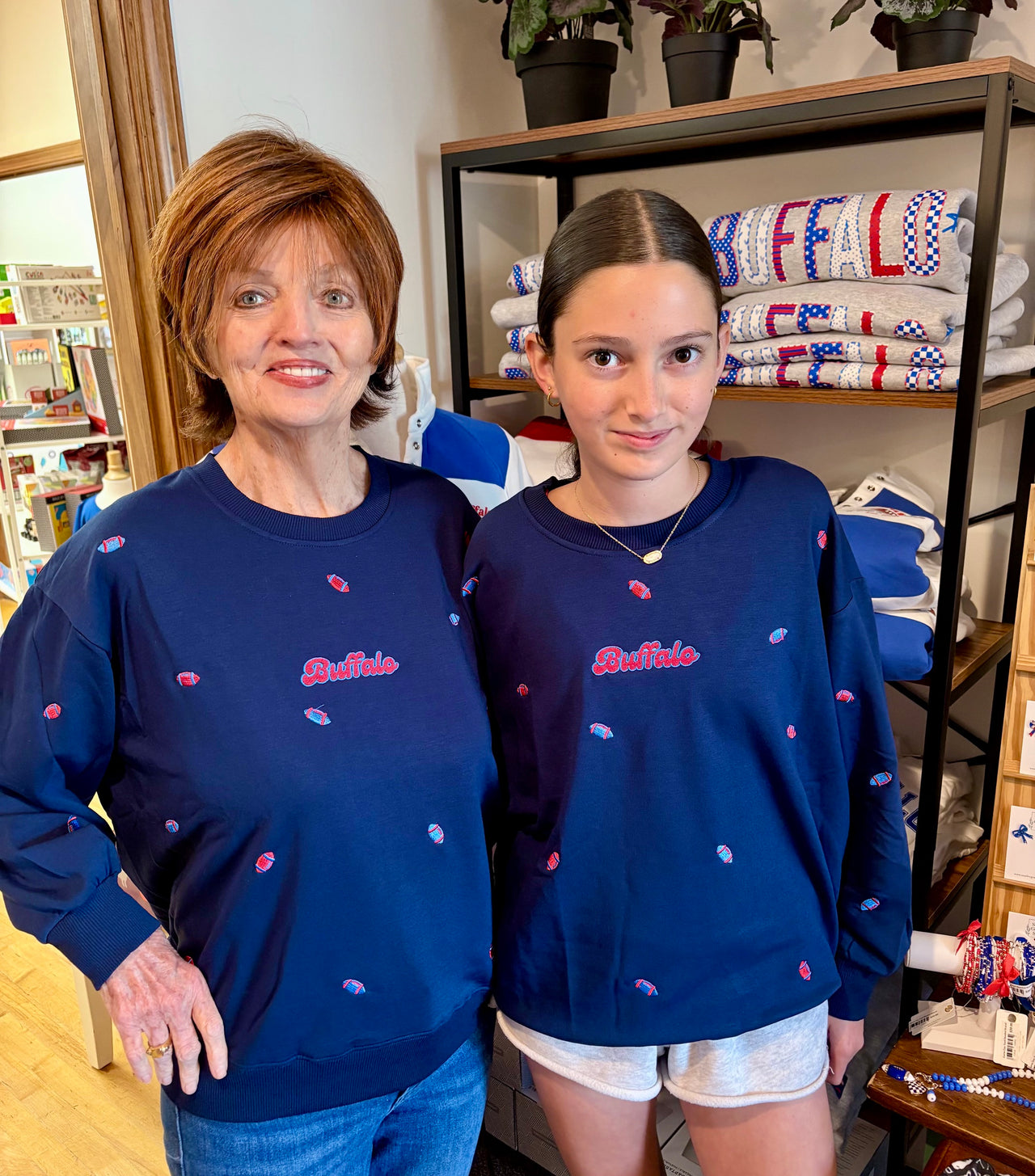 An older woman and a young girl wearing matching navy blue sweatshirts with a red 'Buffalo "go and small red and white embroidered details, standing in a retail store.