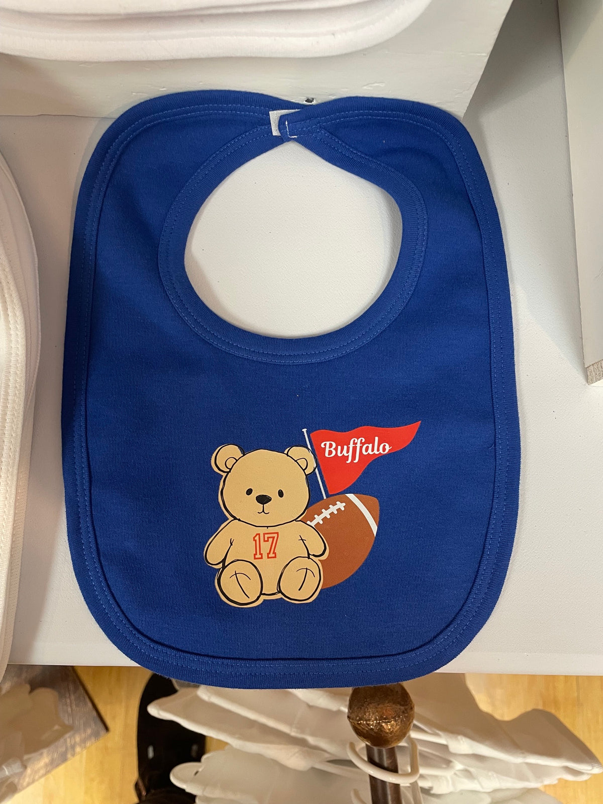 A royal blue baby bib with a custom football-themed graphic featuring a teddy bear, a football, and a red flag that says 'Buffalo'.