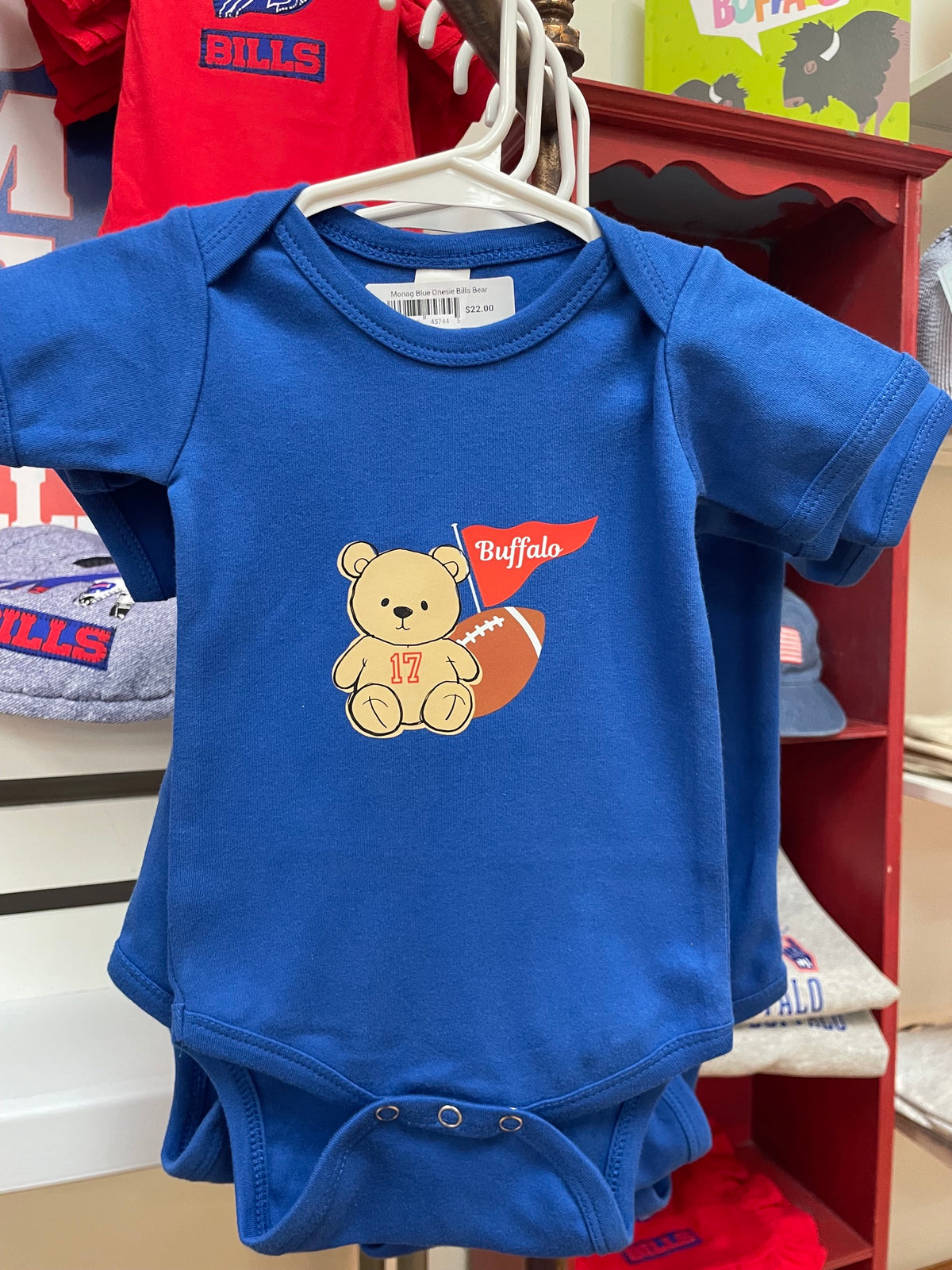 A royal blue baby short-sleeve bodysuit hanging on a white hanger, featuring a cartoon teddy bear holding a football with a red 'Buffalo' flag graphic on the front.